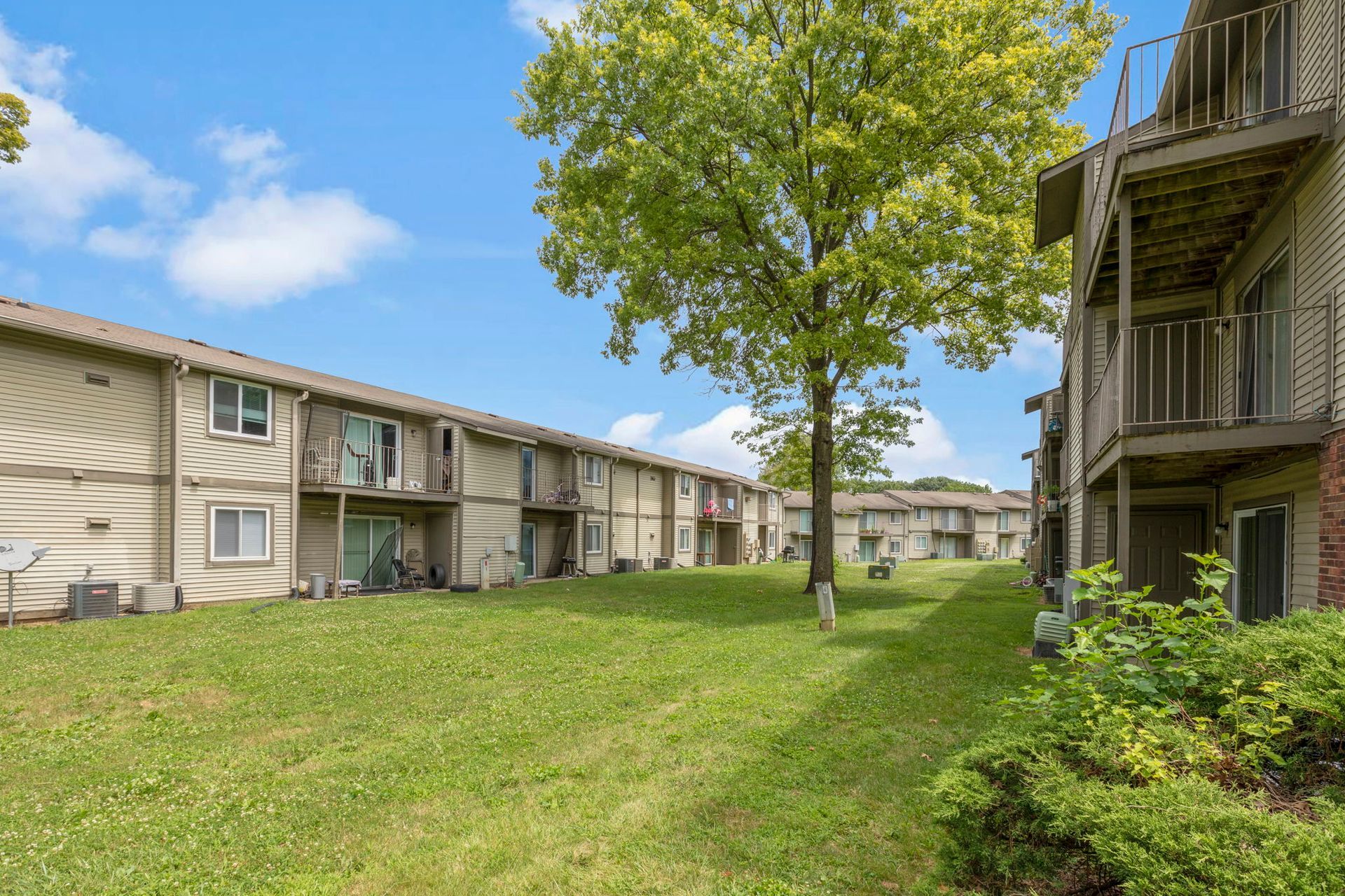 Apartment buildings with balconies overlook a grassy lawn and a tree under a blue sky.