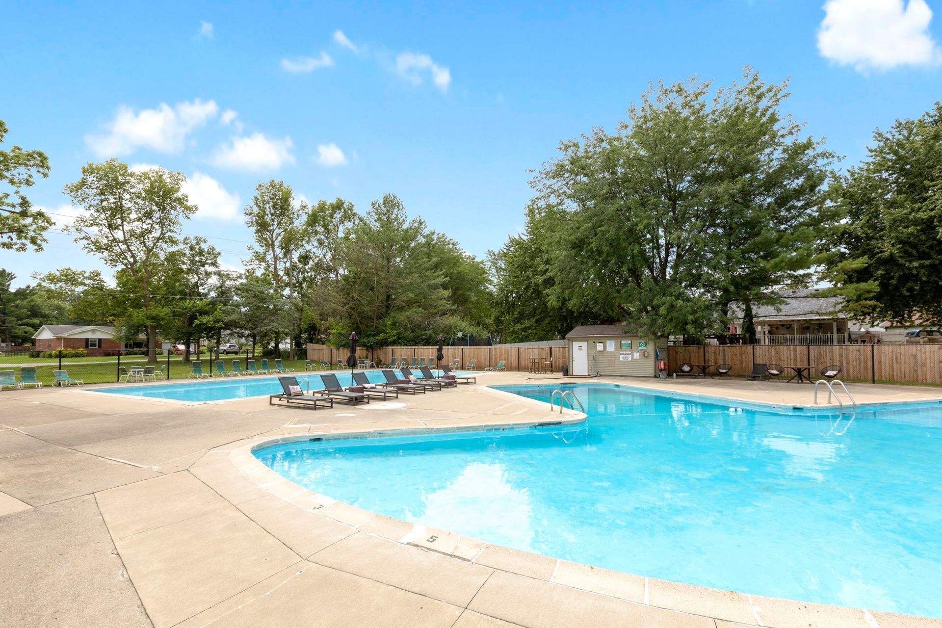 Outdoor swimming pool with lounge chairs, trees, and a building, against a blue sky.