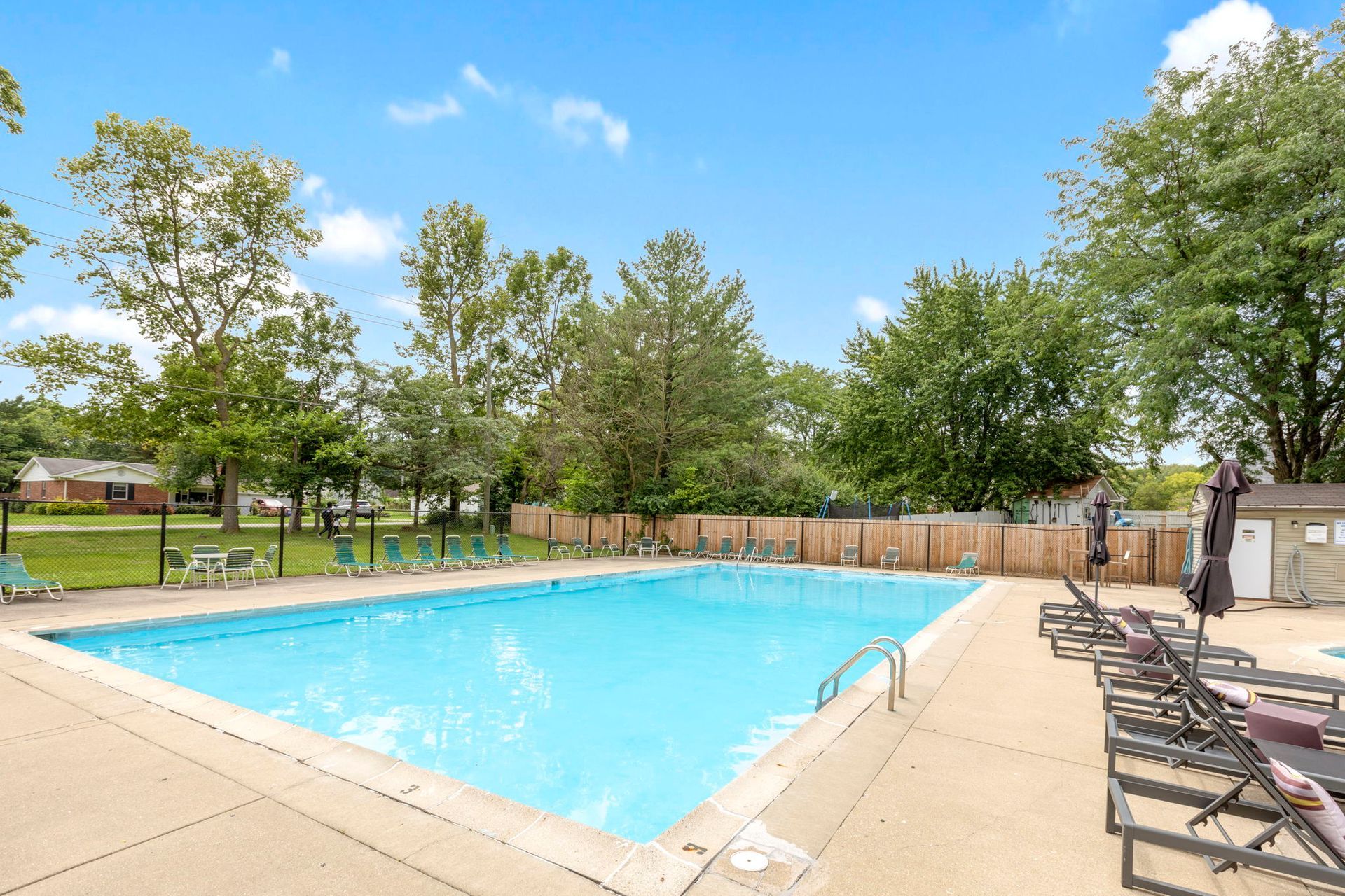 A swimming pool with lounge chairs on a sunny day; surrounded by trees, a fence and small building.