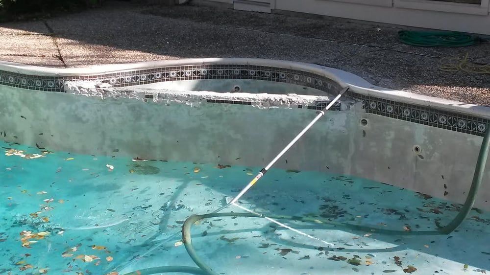 Empty turquoise pool with a white pool vacuum hose and pole inside, beside a concrete edge.