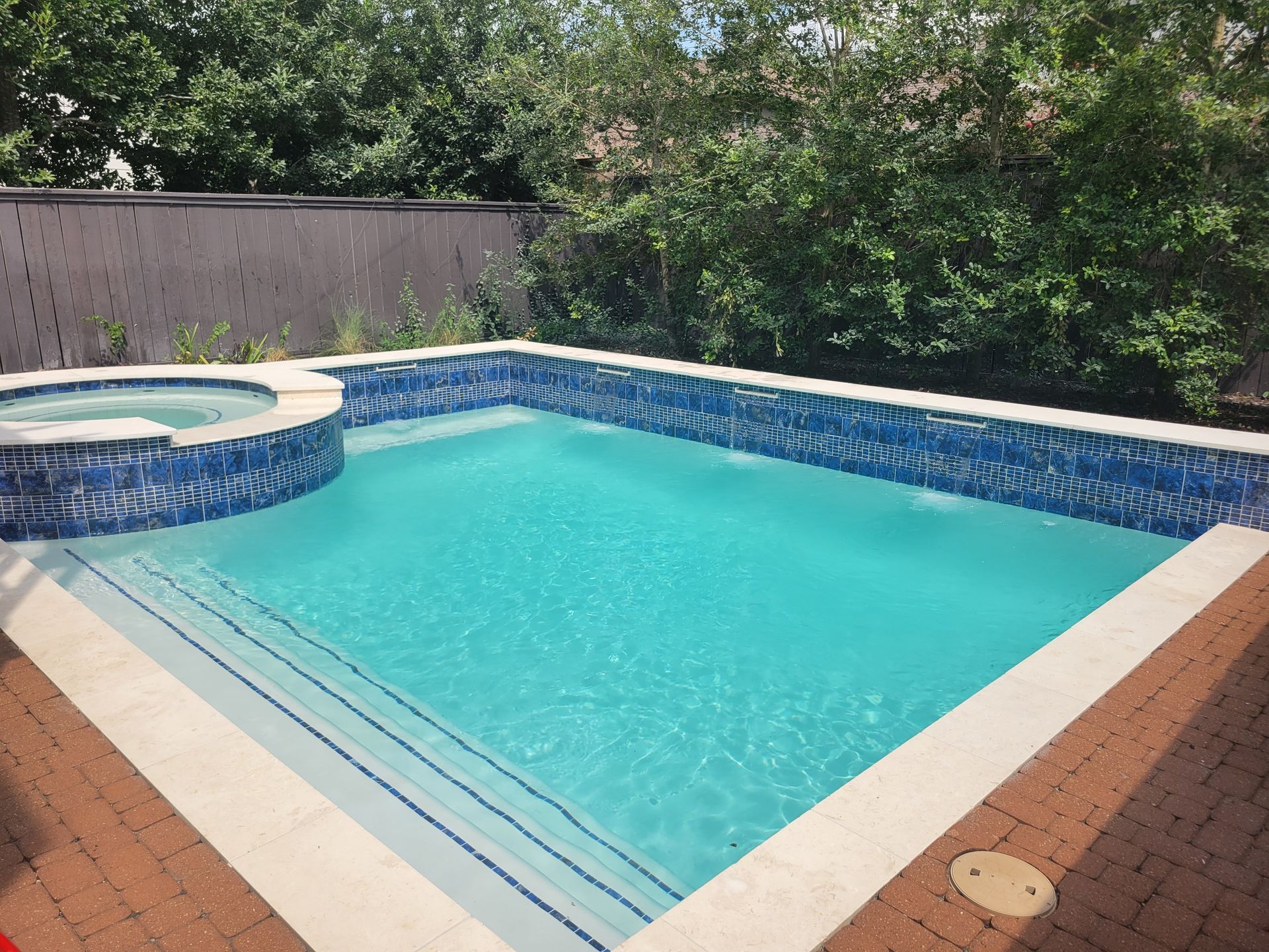 Backyard swimming pool with blue water, brick deck, and spa surrounded by trees and fence