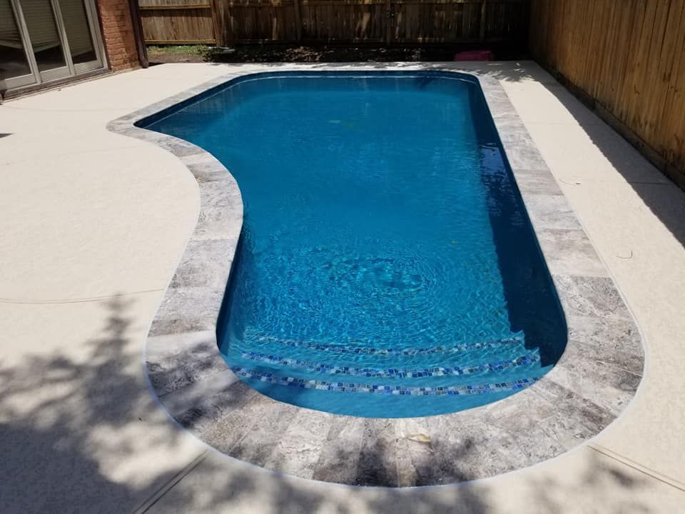 Backyard swimming pool with blue water, curved white stone deck, and wooden fence around it