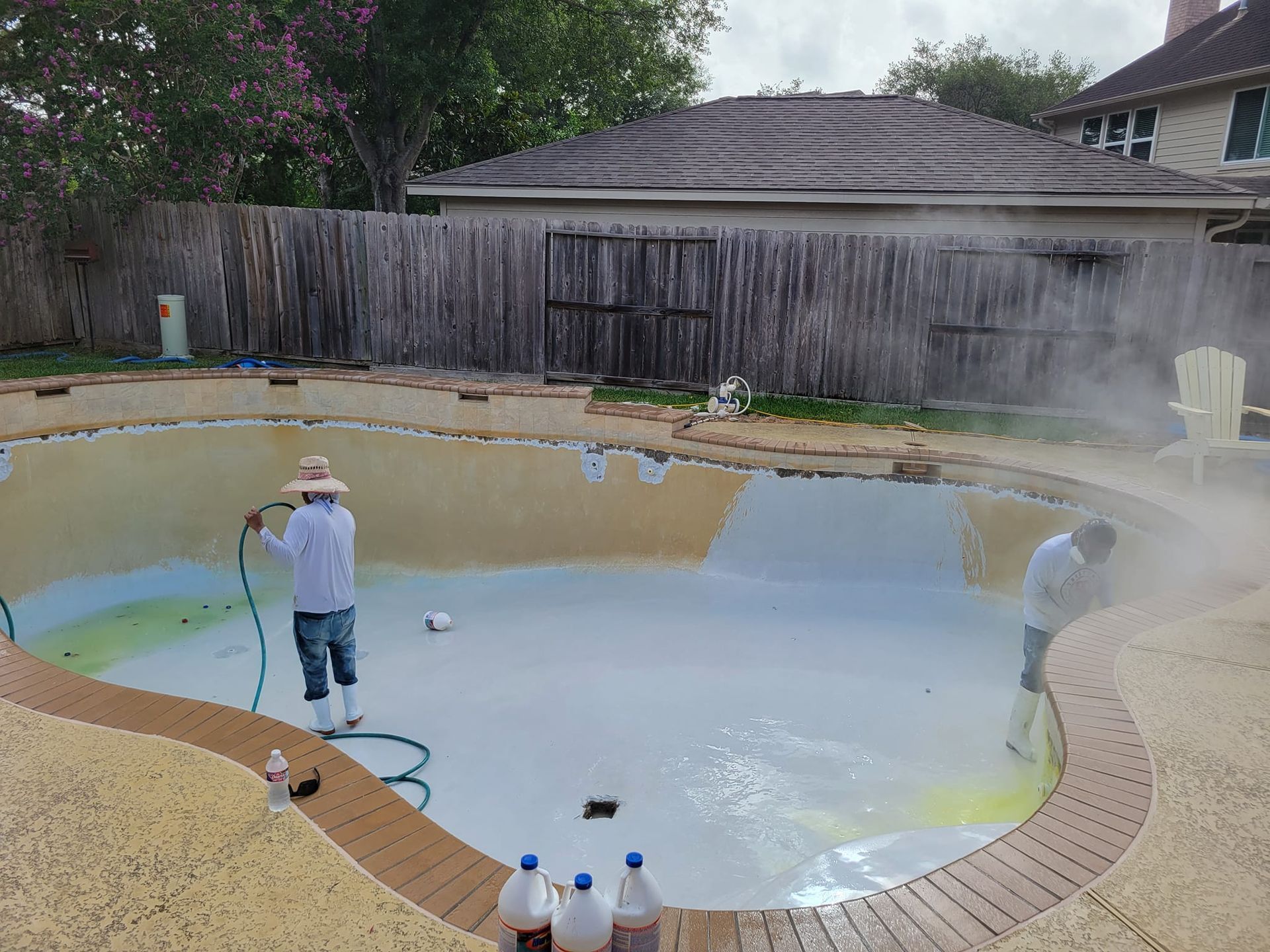 Workers draining a backyard swimming pool with hoses near a fenced yard and house