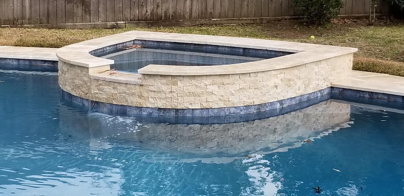 Pool with raised spa and beige stone surround in a backyard.