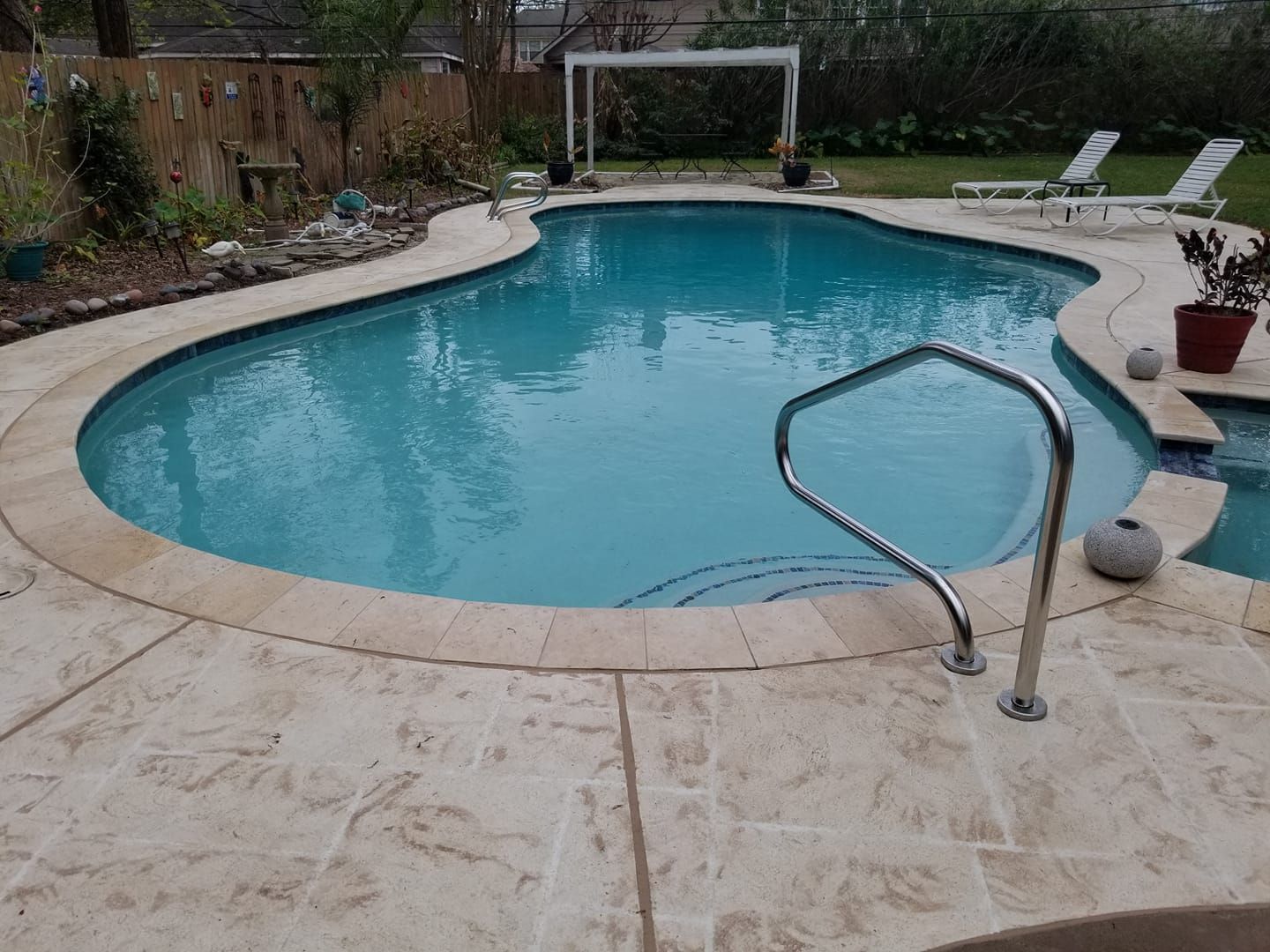 Backyard swimming pool with curved stone deck, handrail, lounge chairs, and green landscaping.