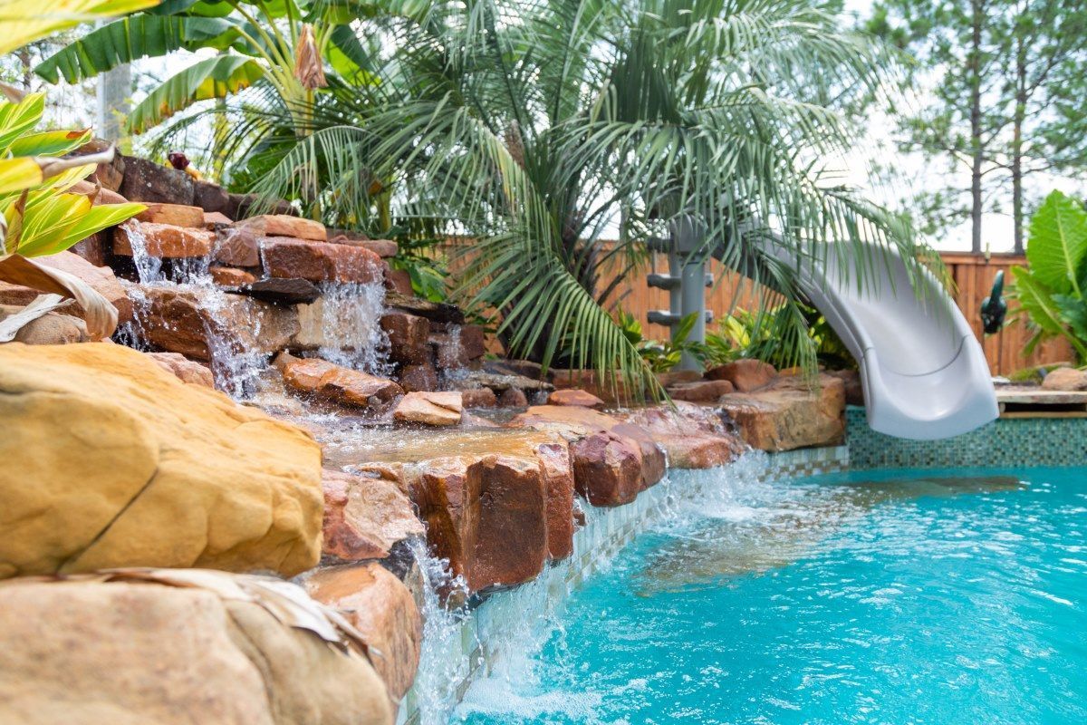 Tropical pool with rock waterfall and silver slide, surrounded by palm trees and tan boulders