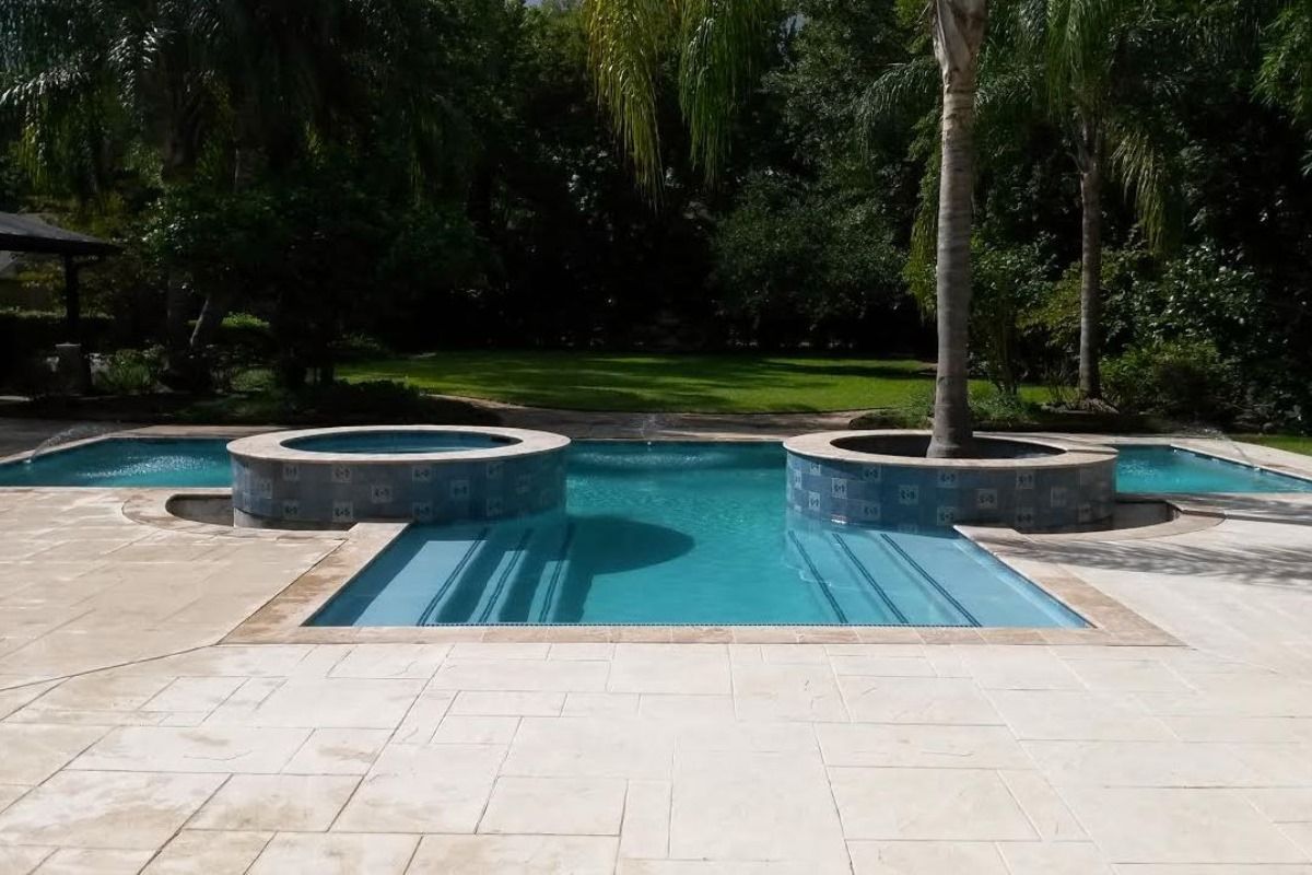Backyard swimming pool with blue water, stone deck, and two round spa features among palm trees