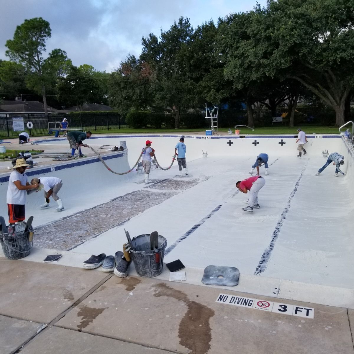 Workers cleaning an empty outdoor swimming pool with hoses and tools, trees in the background.