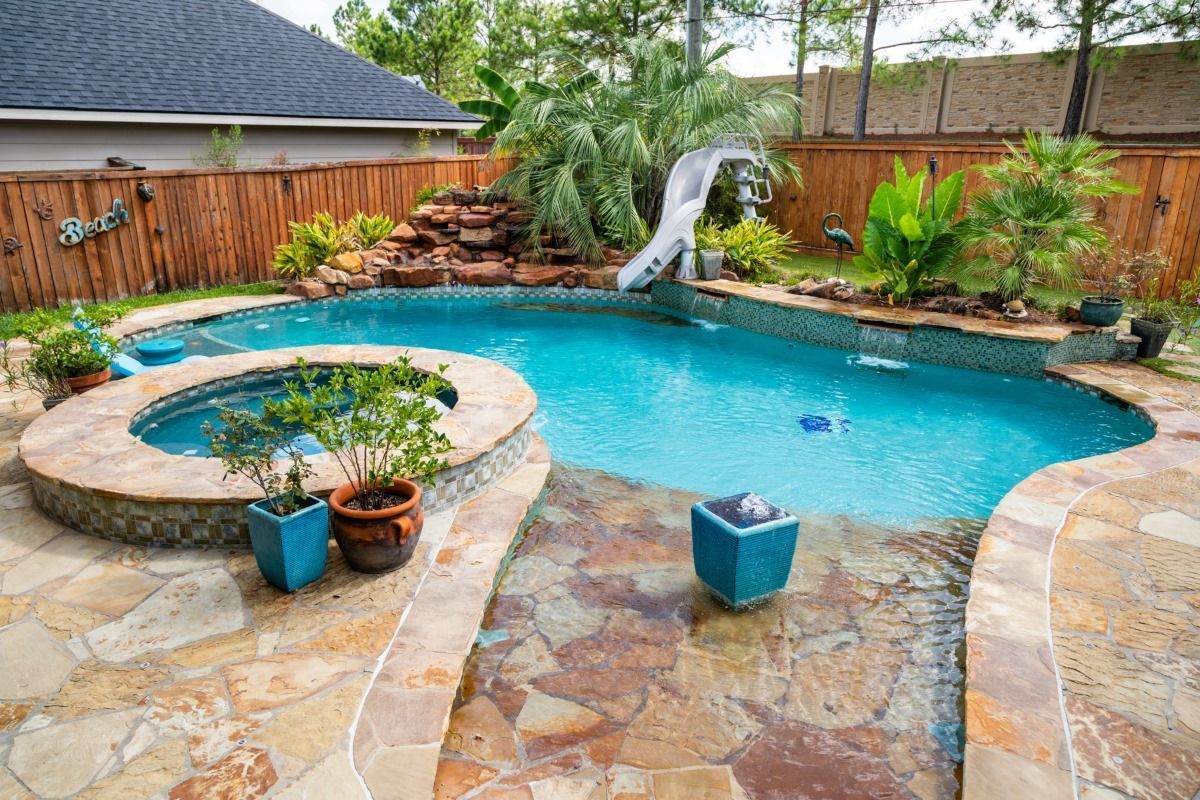 Backyard pool and spa with stone patio, turquoise water, potted plants, and a small waterfall slide