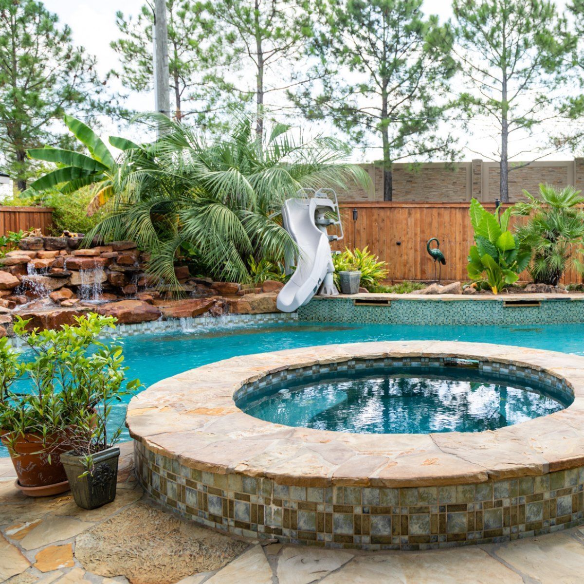 Backyard pool with circular hot tub, slide, waterfall rocks, and wooden fence