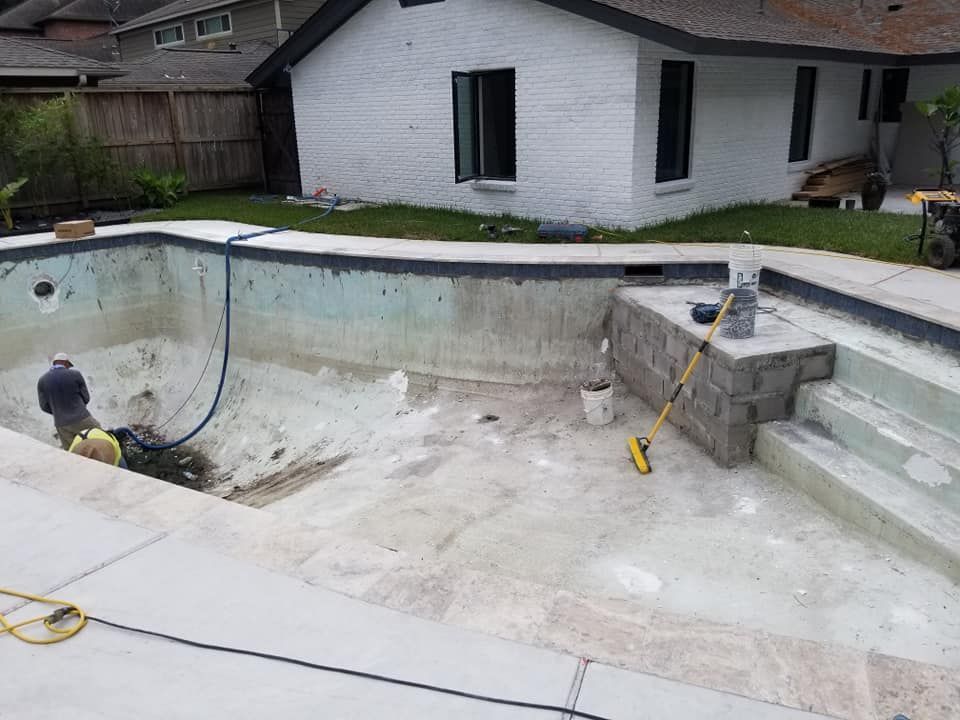Empty drained backyard pool under renovation beside a white house, with tools and concrete steps visible