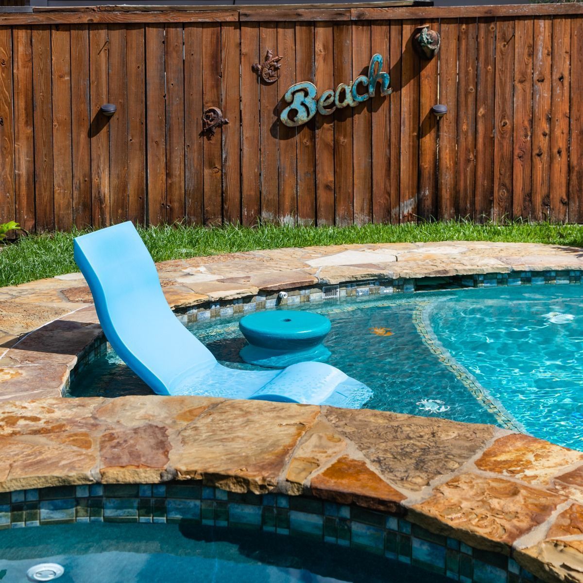 Backyard pool with a blue in-pool lounge chair, round side table, stone edging, and wooden fence