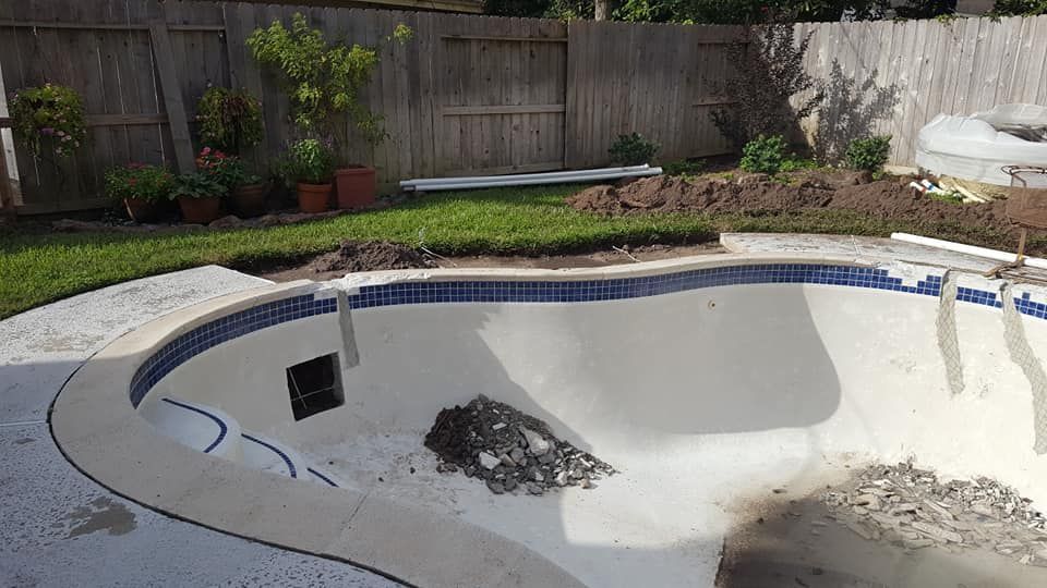 Empty swimming pool with a large crack in the floor and debris inside, in a backyard with a fence and plants