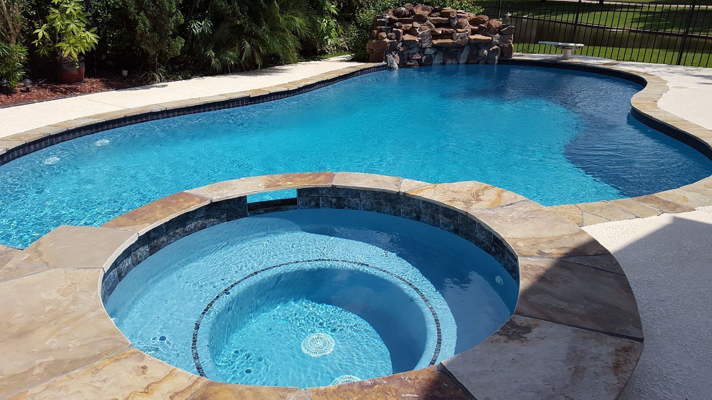 Backyard pool with a raised circular hot tub and blue water, framed by stone decking and a waterfall backdrop