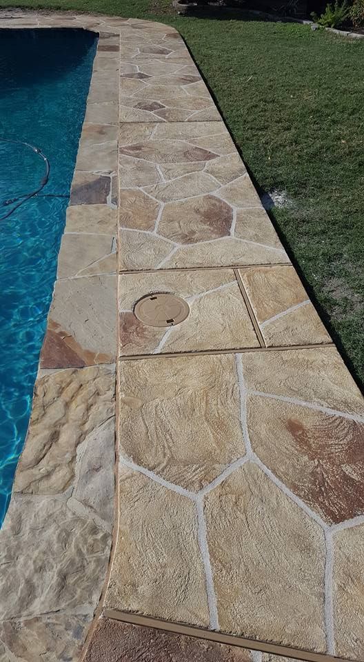 Stone pool deck beside blue swimming pool and green lawn, with a circular drain cover in the paving.