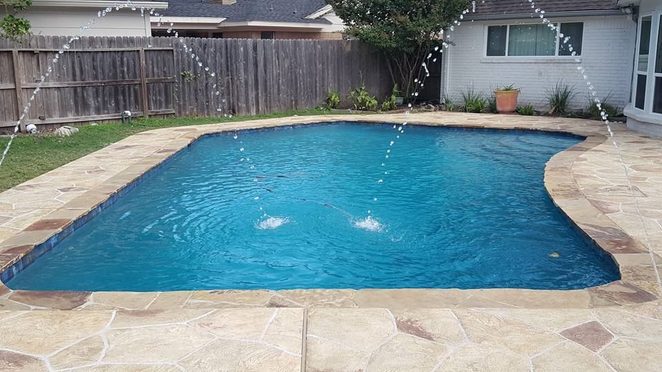 Backyard swimming pool with blue water, surrounded by stone patio and wooden fence.