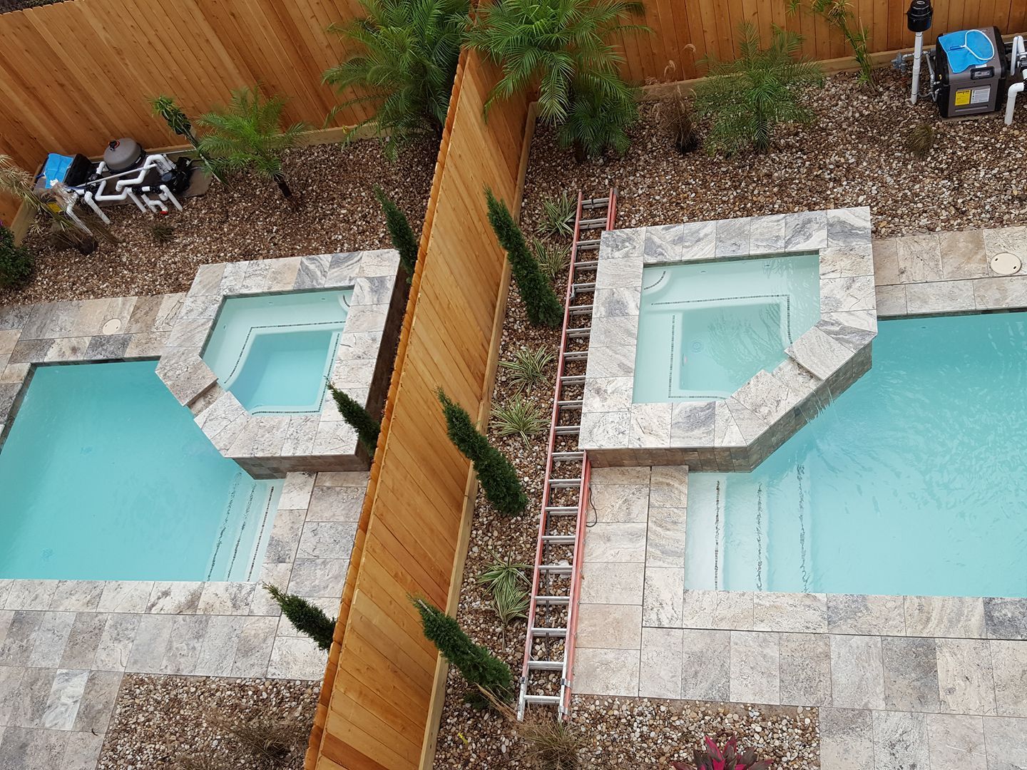 Overhead view of two backyard pools separated by a wooden fence, with stone patios and a ladder on the right.