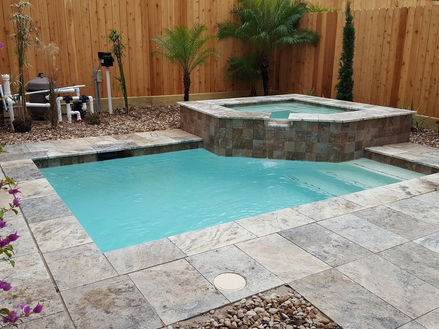 Backyard pool with turquoise water, stone patio, and raised spa beside a wooden fence