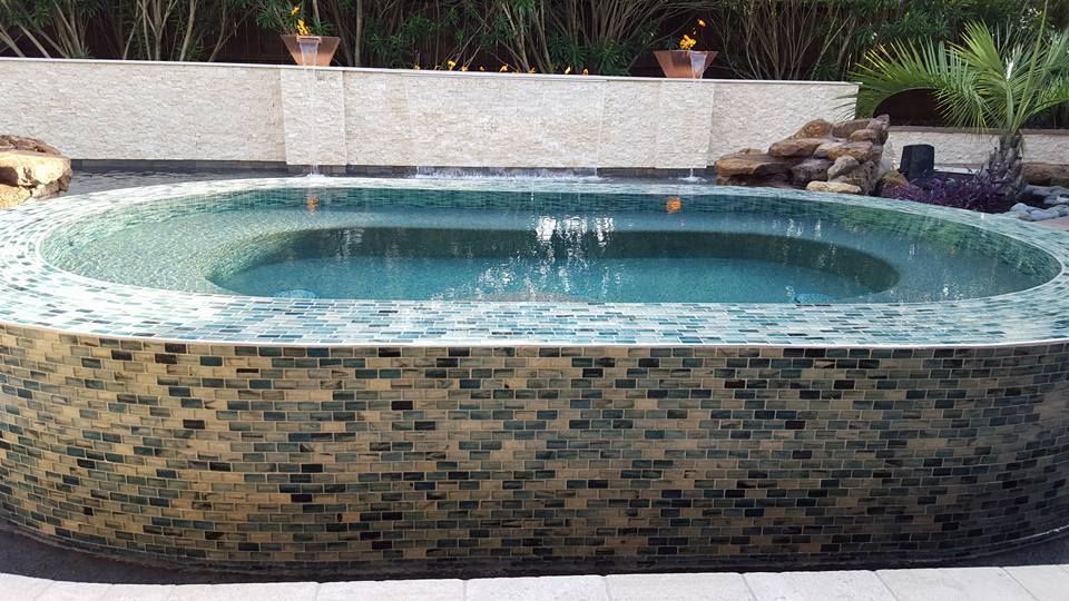 Small circular backyard pool with blue water and mosaic tile edging, set among greenery.