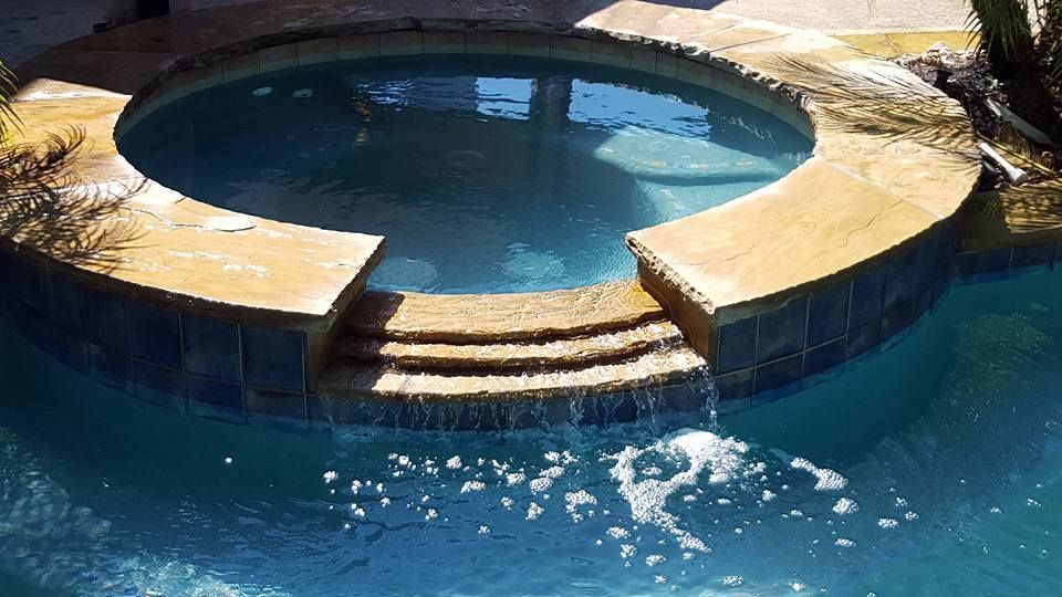 Round stone hot tub with blue water and steps, bubbling beside a larger pool.