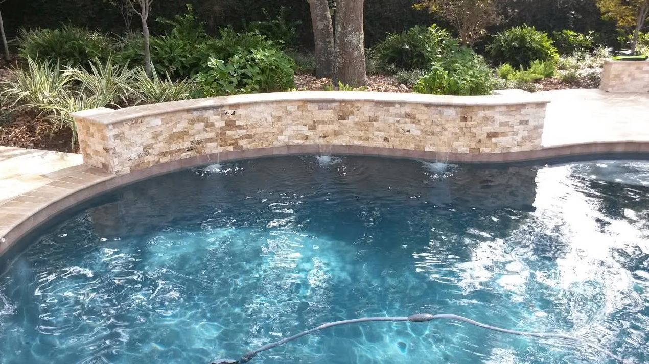 Backyard swimming pool with dark blue water and a curved stone retaining wall surrounded by greenery