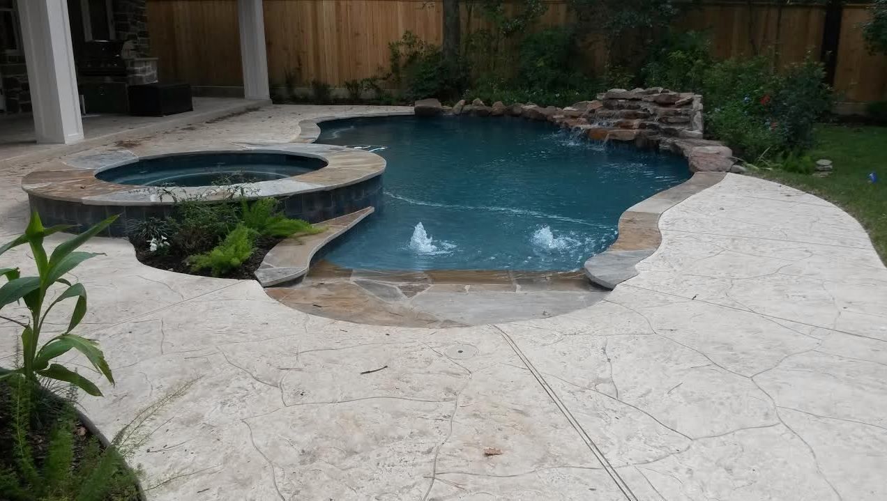 Backyard swimming pool with stone spa, light patio, and wooden fence surrounded by greenery