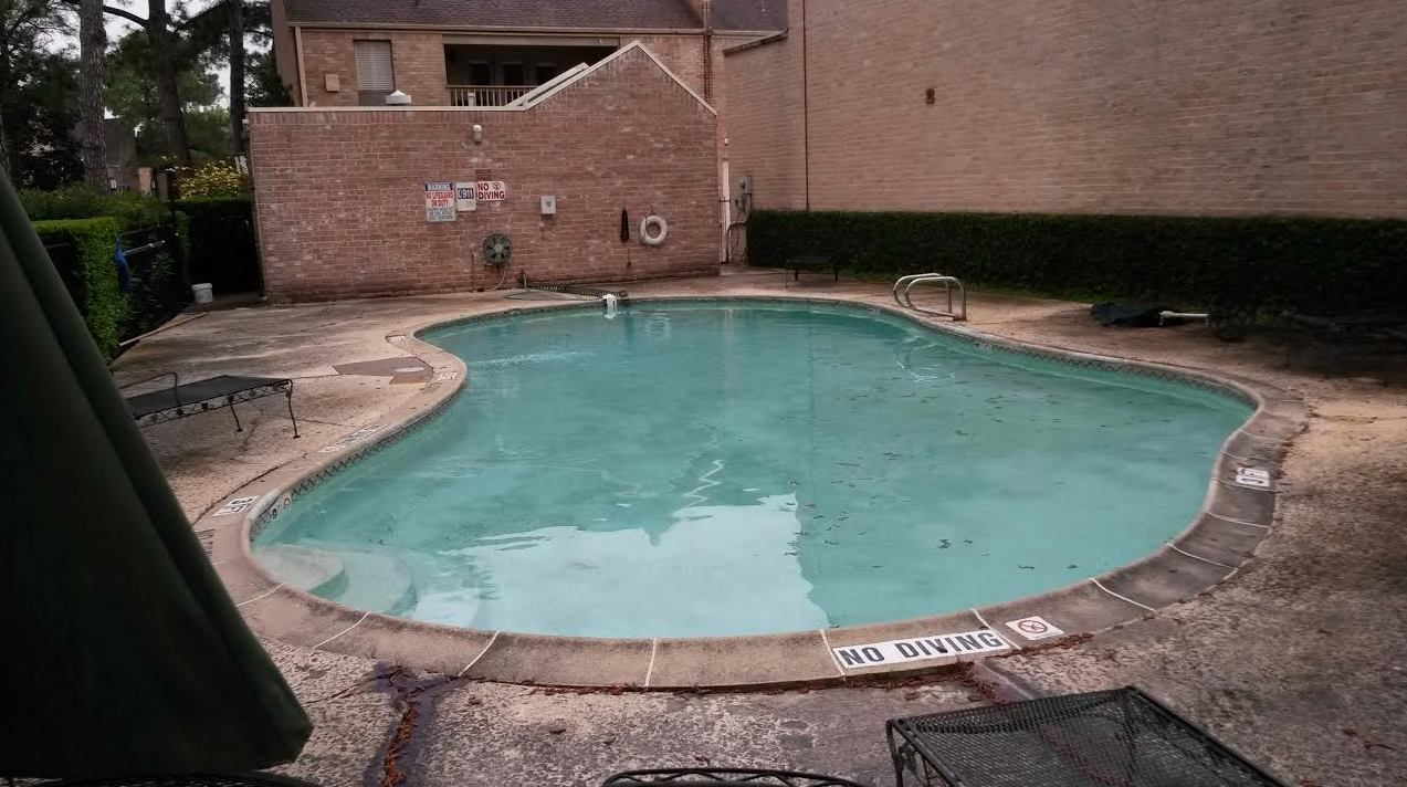 Small outdoor swimming pool with turquoise water, surrounded by a brick patio and apartment buildings.