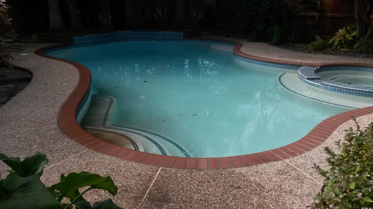 Curved backyard swimming pool with blue water and pebble deck at night