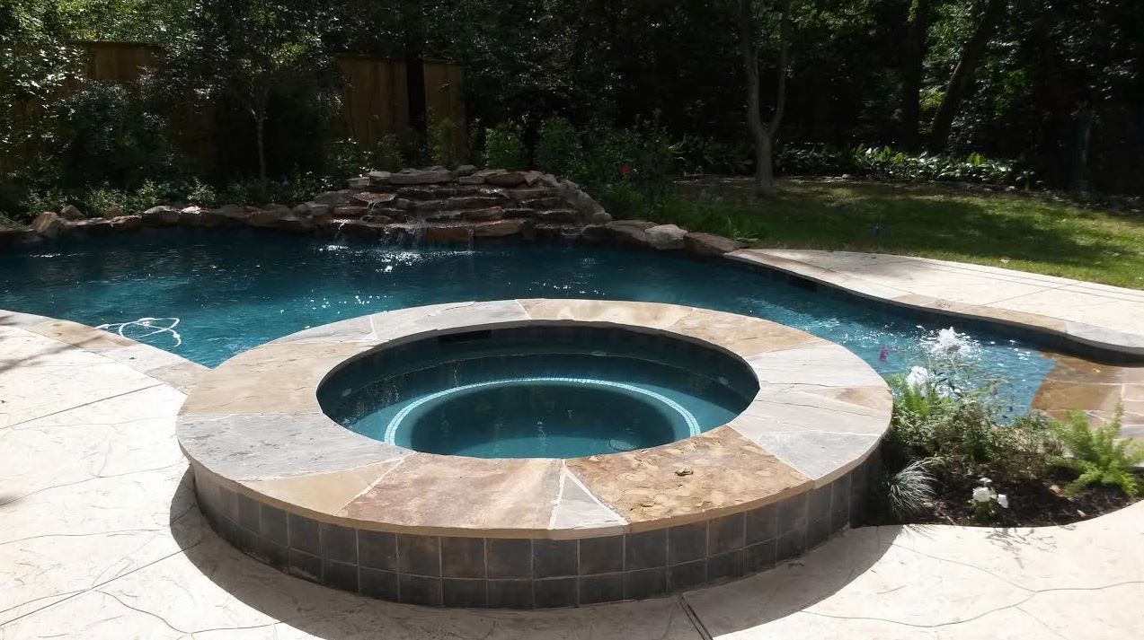 Backyard pool with a circular stone hot tub and surrounding patio in bright sunlight