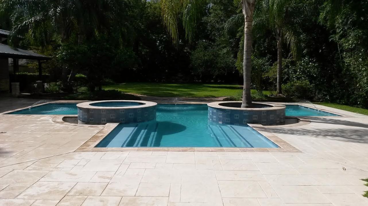 Backyard pool with a central walkway, two round hot tubs, and surrounding patio in a wooded setting