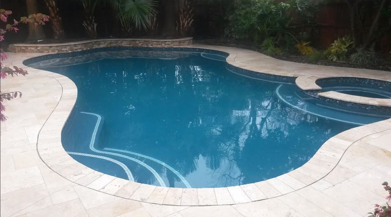 Curved backyard swimming pool with blue water and light stone decking, surrounded by greenery