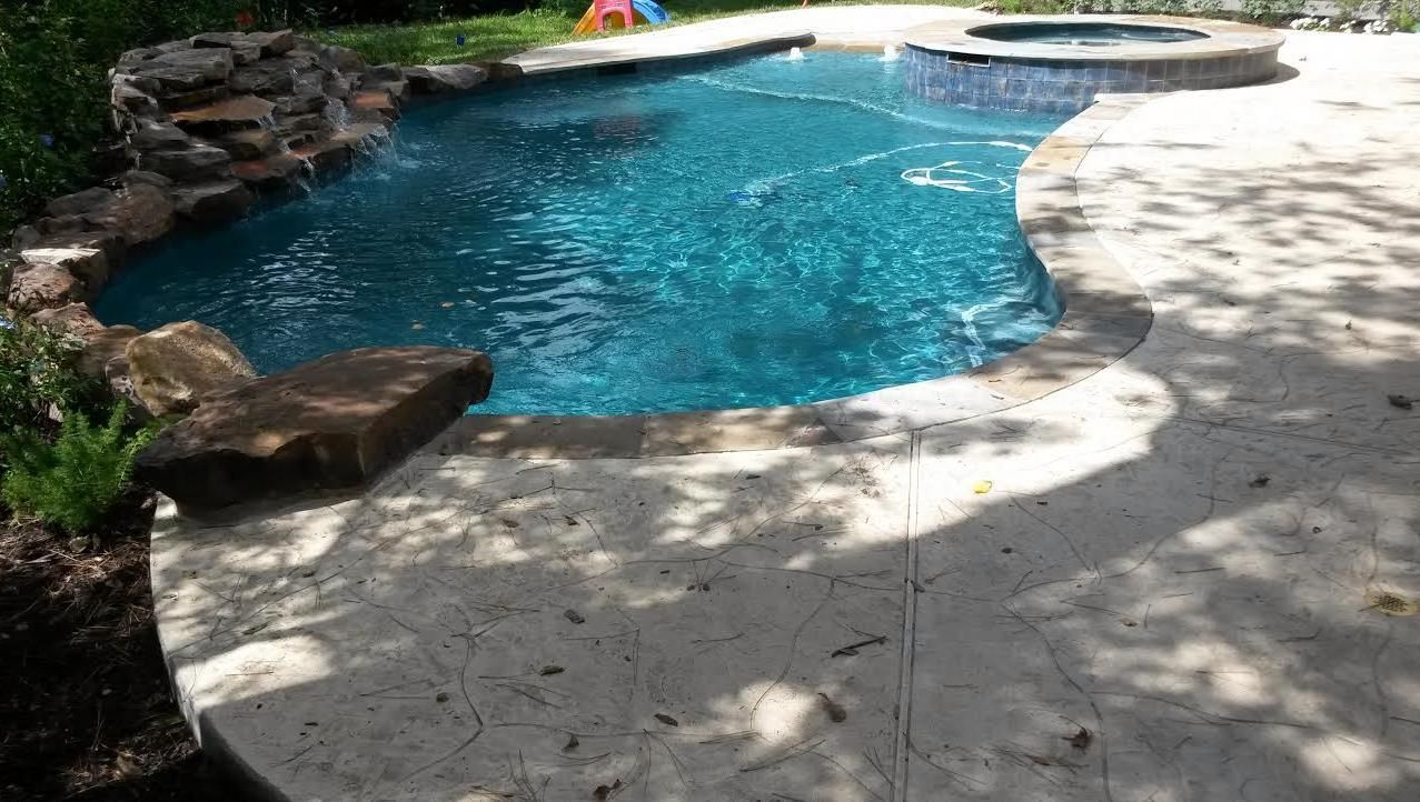 A blue backyard pool with stone edging and a sunlit patio around it.