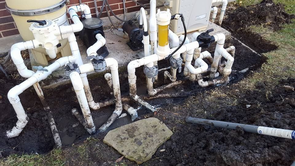 White PVC pipes and valves in a muddy outdoor utility area beside tanks and soil.