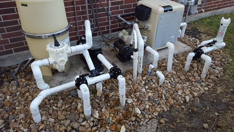 White utility pipes and valves beside a house on gravel, with a tan tank and beige equipment unit.