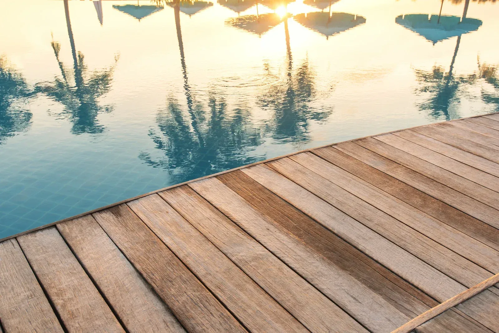 Wooden deck next to a pool reflecting palm trees and umbrellas at sunset.