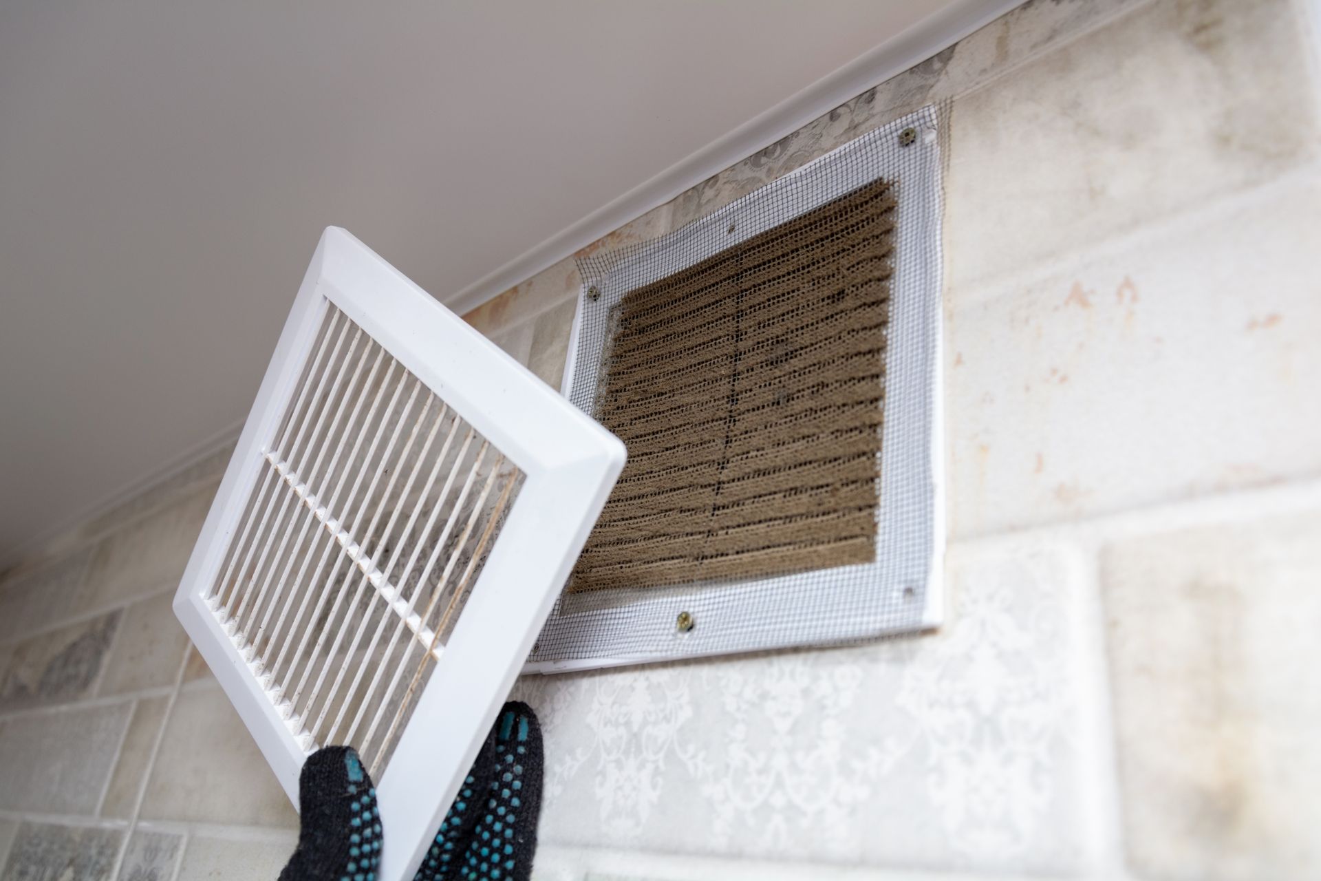 Gloved hands removing a dusty vent cover from a wall air duct grille during cleaning