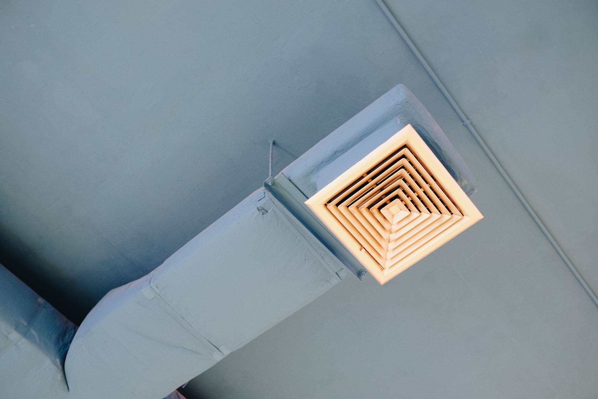 A ceiling air vent connected to a rectangular duct painted in light gray. A ceiling air vent connected to a rectangular duct painted in light gray.