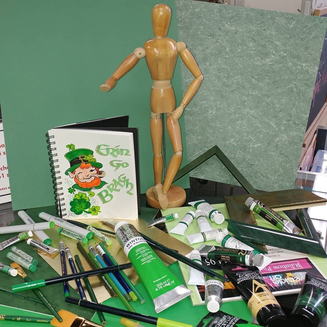 Wooden artist's mannequin with green art supplies: paints, pencils, and a sketchbook with a leprechaun.