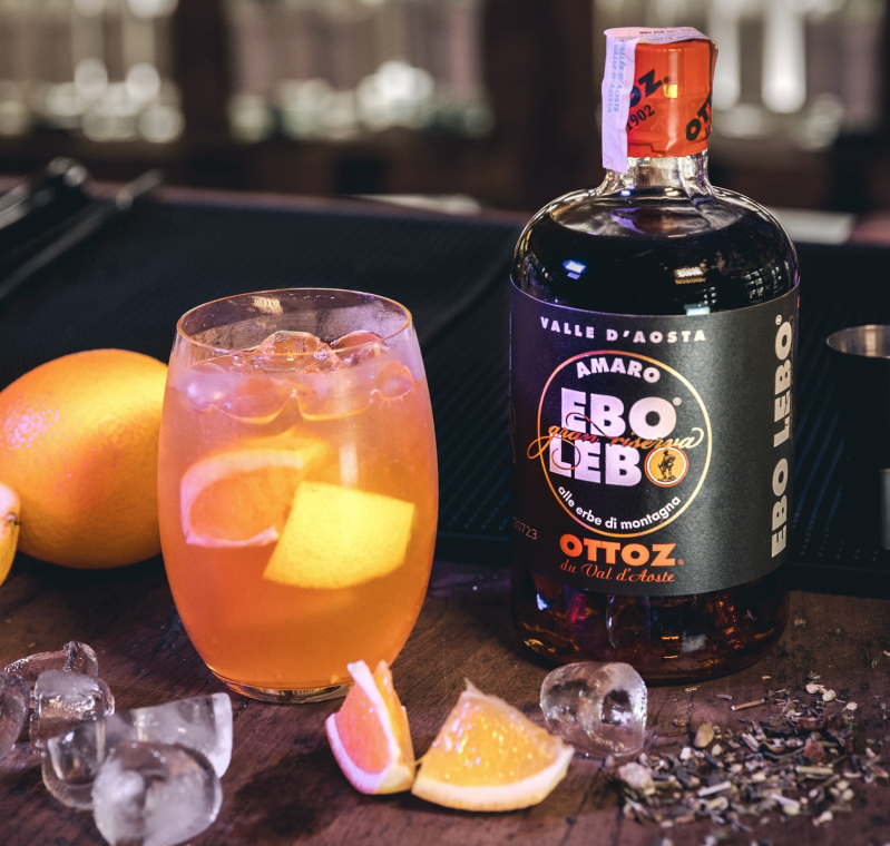 Cocktail con ghiaccio e guarnizione d'arancia accanto a una bottiglia di Amaro e arance su un piano di bar scuro.