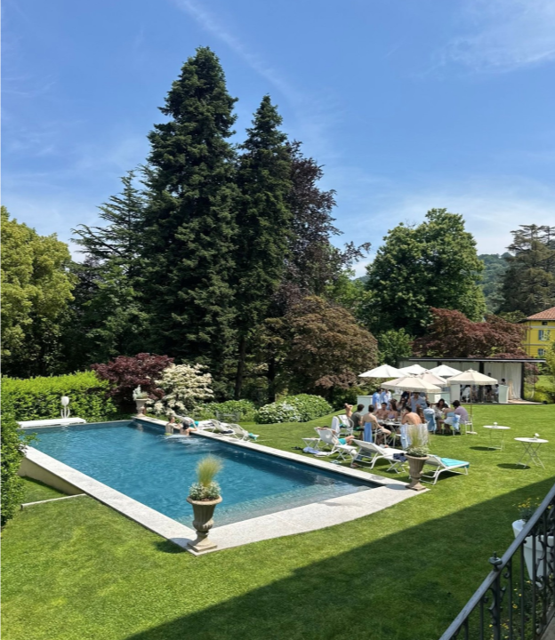 Piscina in giardino con lettini, ombrelloni e persone su un prato soleggiato circondato da alberi e montagne.