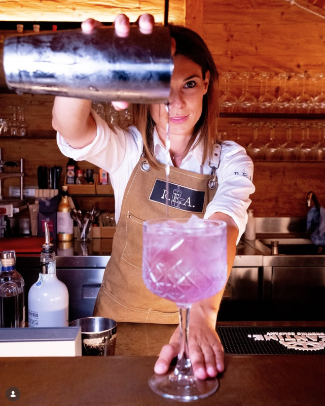 Un barista seduto su un bancone di legno versa un cocktail rosa in un calice di vetro.