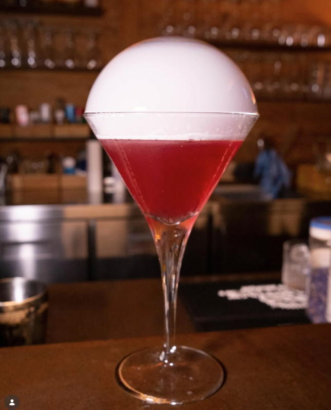 Cocktail rosso in un bicchiere con stelo sormontato da una cupola di schiuma bianca, servito in un bar.