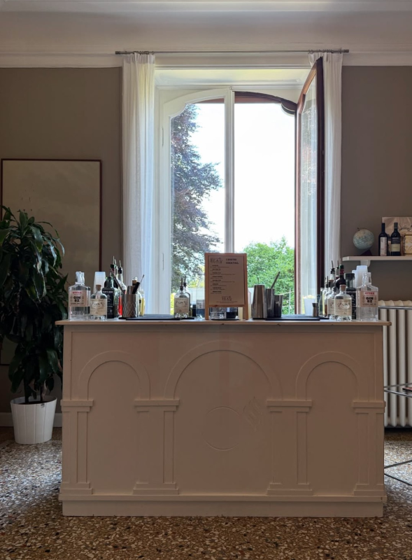 Bancone bar bianco in una stanza, con bottiglie e oggetti decorativi davanti a una finestra aperta che si affaccia sugli alberi.