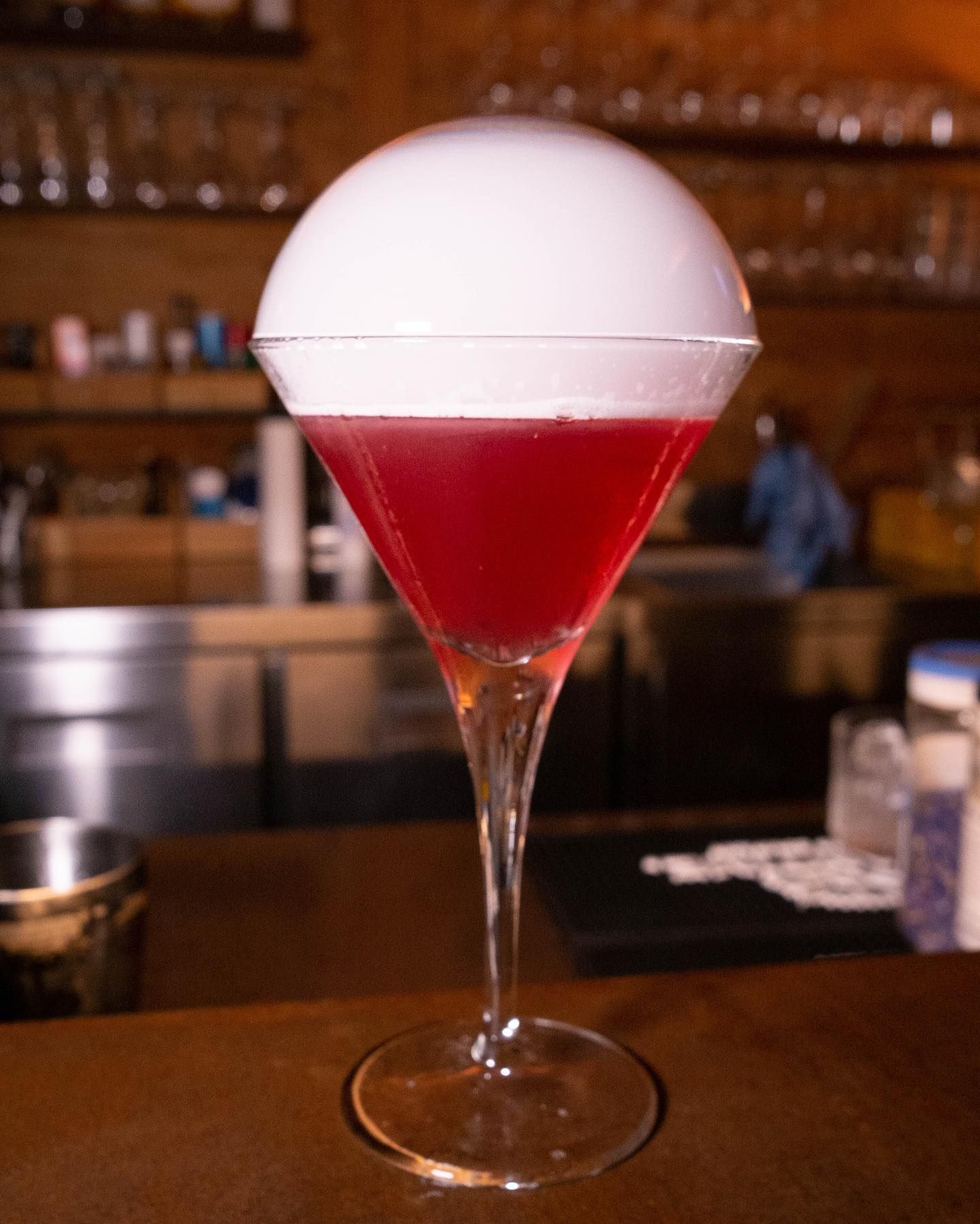 Cocktail rosso in un bicchiere con stelo sormontato da una cupola bianca, su un bancone da bar.