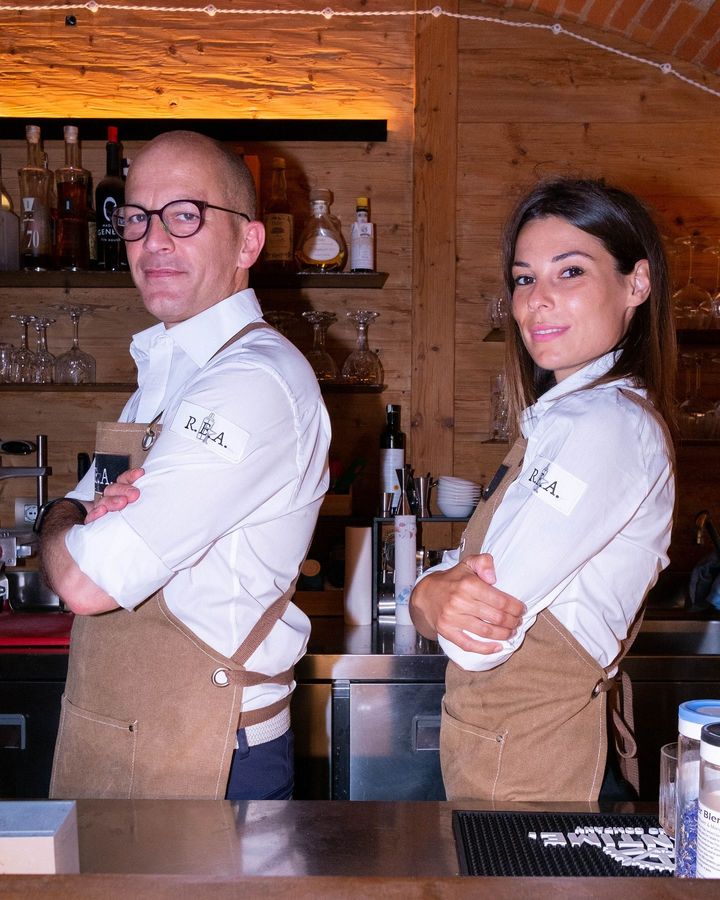 Due baristi in camicia bianca e grembiule in piedi dietro un bancone di legno con le braccia incrociate