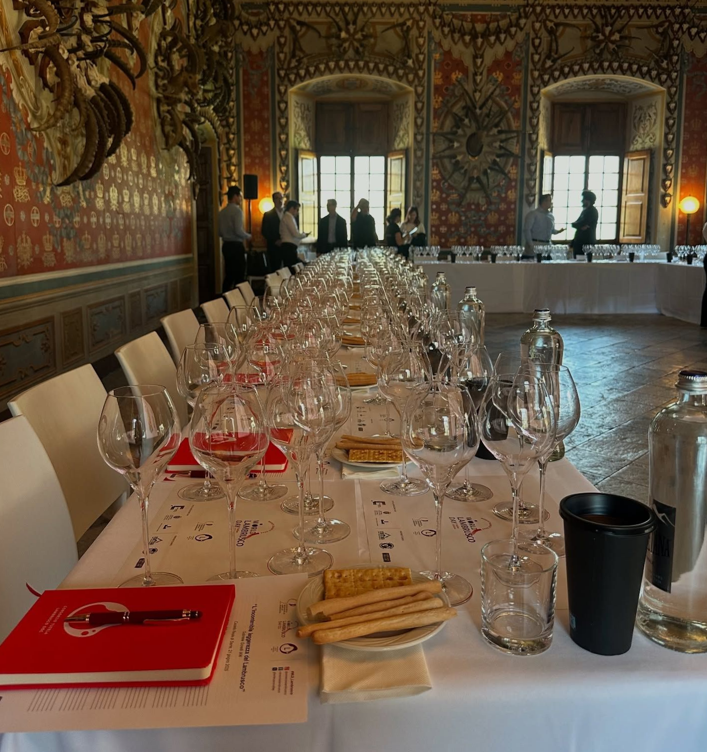 Un lungo tavolo da banchetto apparecchiato con calici da vino e posate in una sala riccamente decorata.