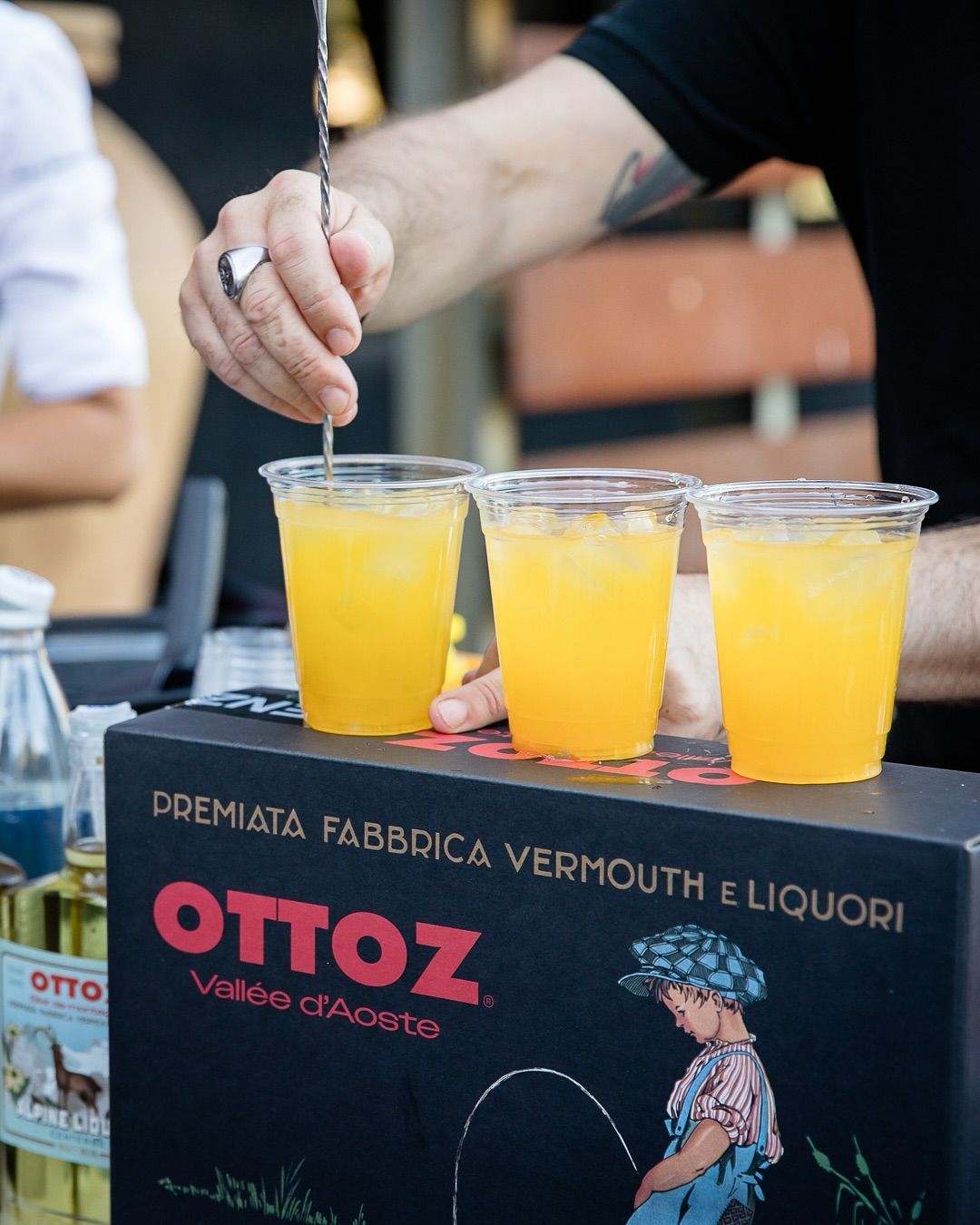 Un barista mescola bevande di un arancione brillante in bicchieri di plastica su un tavolo espositivo di vermouth OTTOZ.
