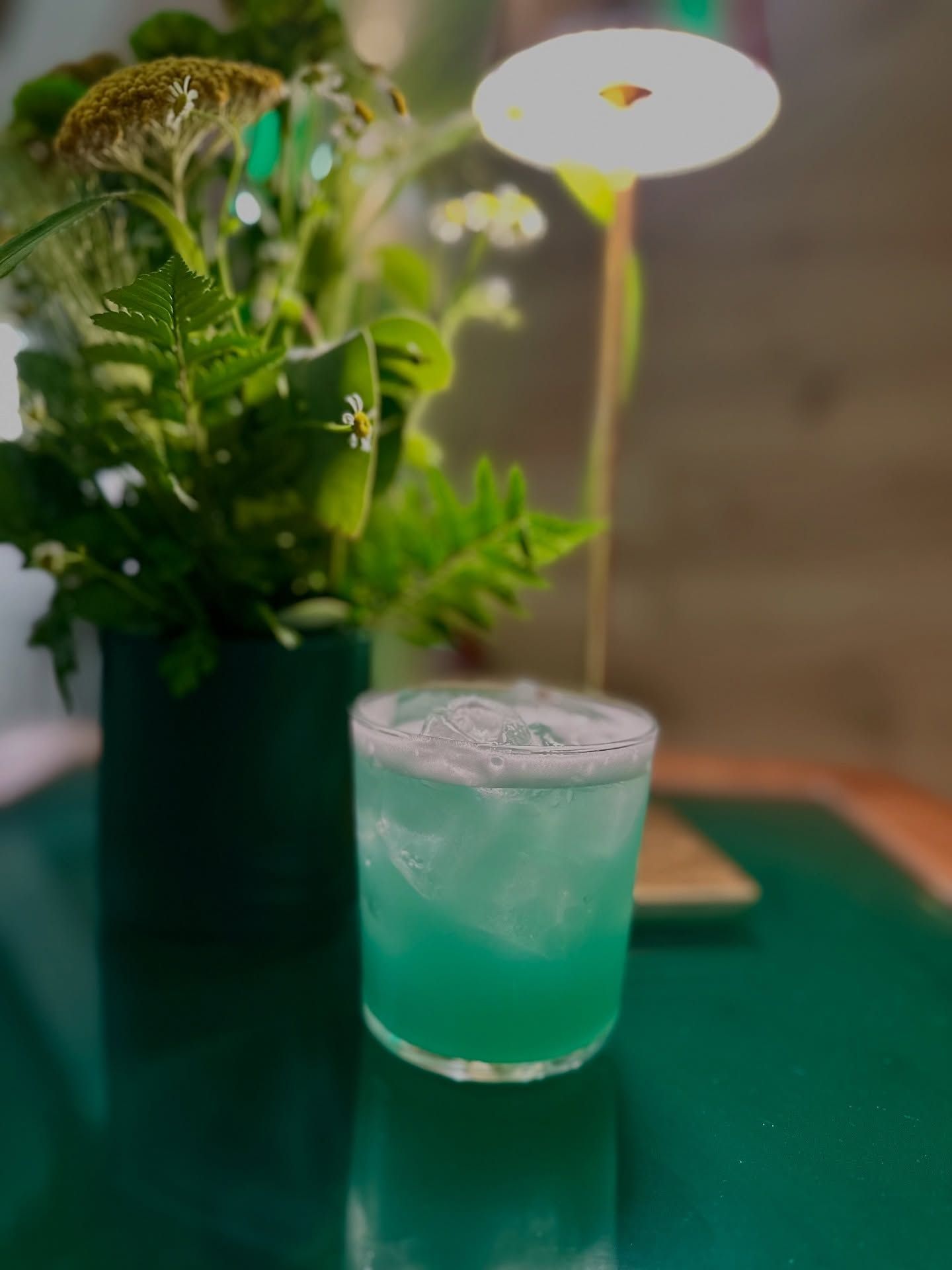Cocktail verde con ghiaccio accanto a una composizione floreale su un tavolo con una piccola lampada