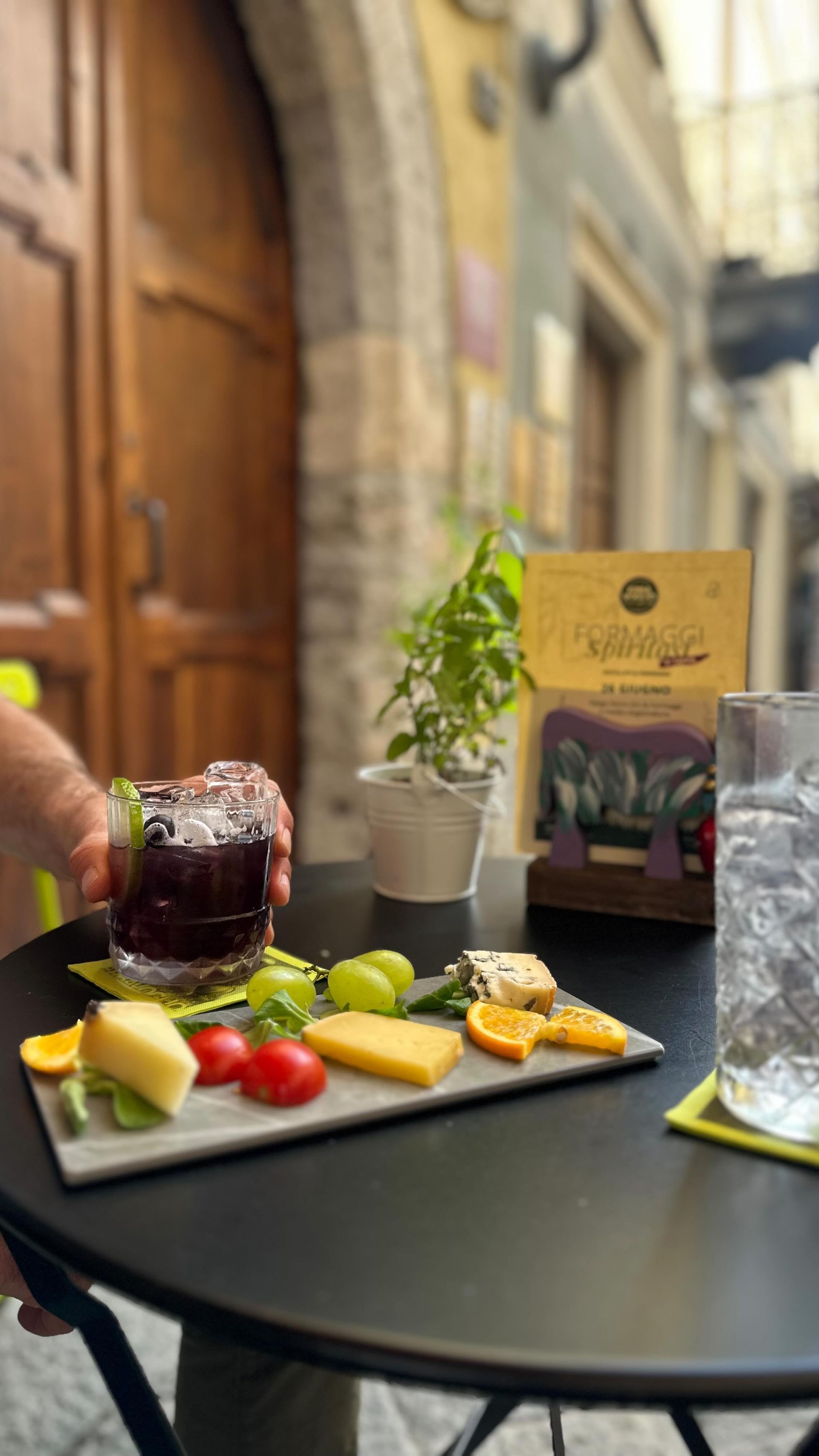 Tavolo all'aperto con frutta a fette, formaggio, vino e una pianta in vaso accanto a un menù giallo.