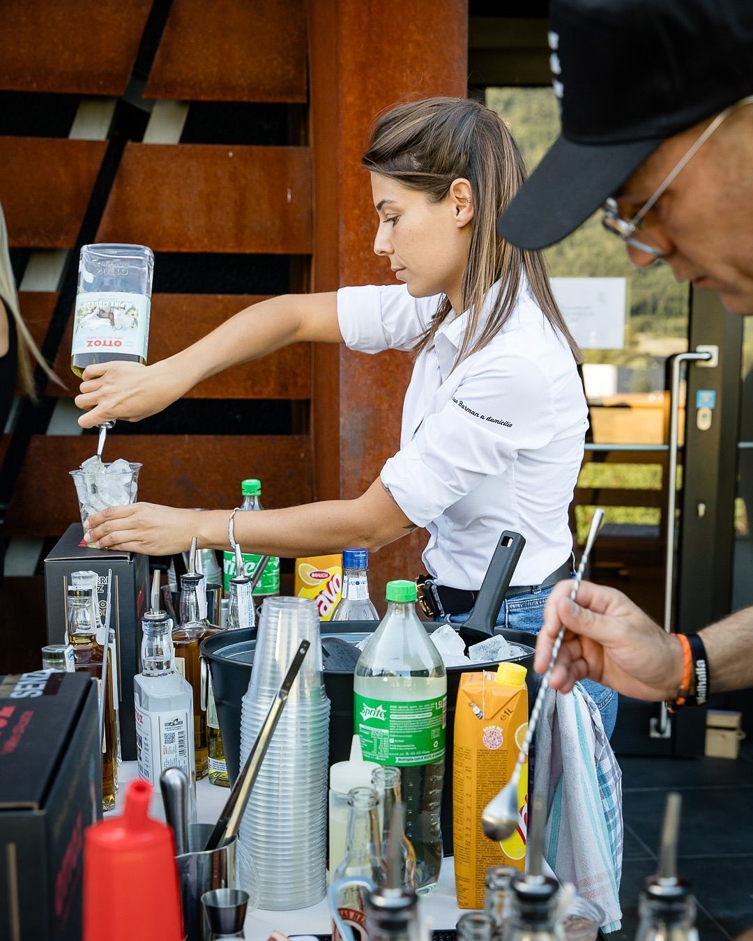 Un barista versa da bere in un bar affollato, con bottiglie, mixer e bicchieri in primo piano.